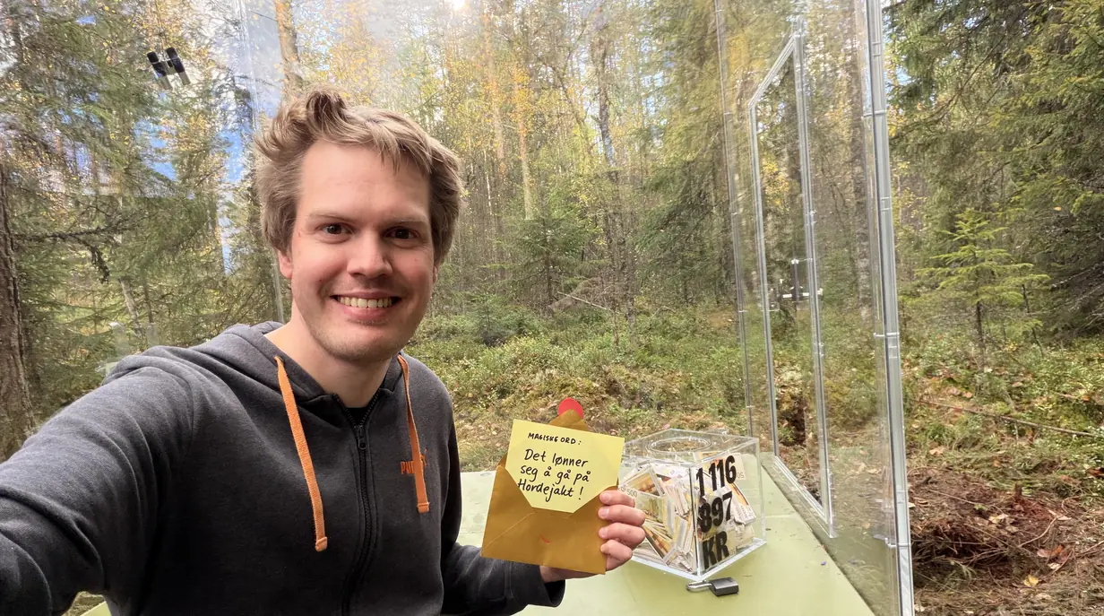 En smilende finner holder opp konvolutten med de magiske ordene ved siden av pengekassen i skogen.