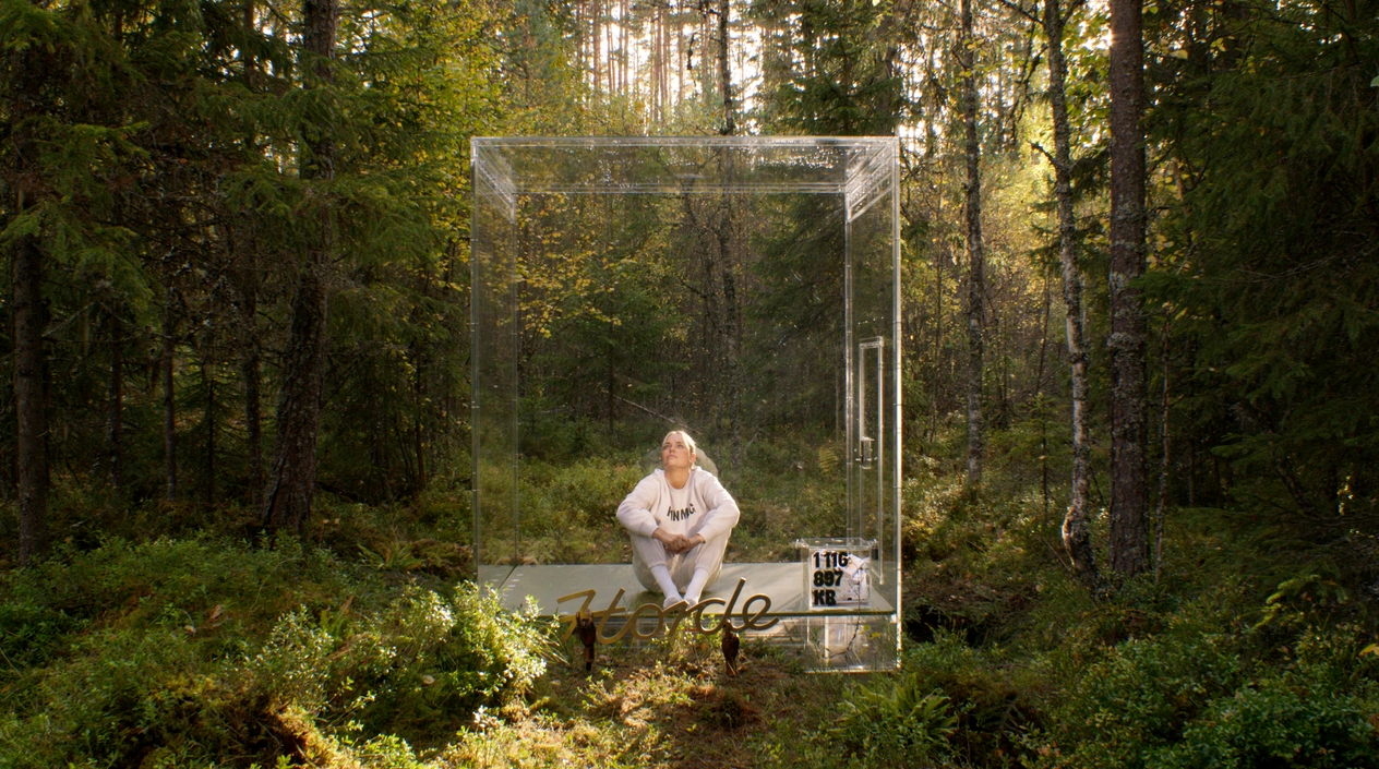 Den transparente pengekassen står i skogen, med en person inne i den.