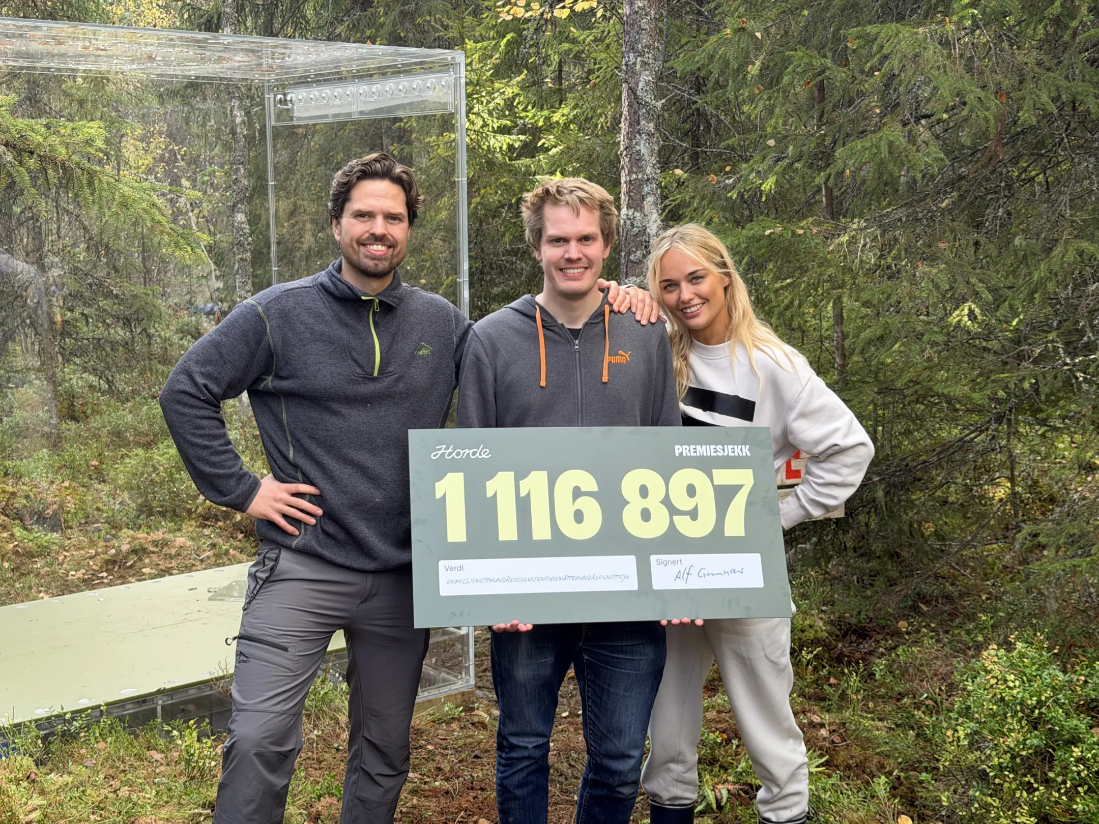 Knut holder premiesjekken på 1 116 897 kroner, sammen med Anja og Alf, foran den gjennomsiktige kassen i skogen.